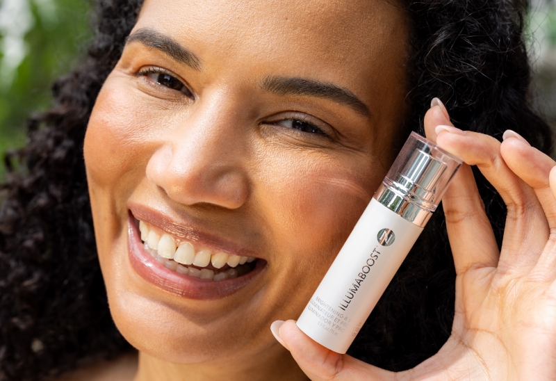 Image of woman smiling, holding bottle  of IllumaBoost Vitamin C Serum