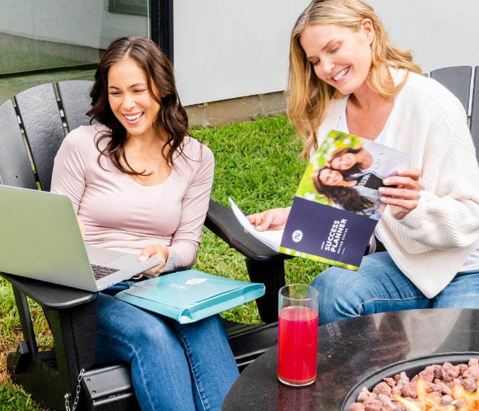 Photograph of two women sitting together around a backyard firepit, smiling and looking at a computer laptop and through Neora opportunity brochures.