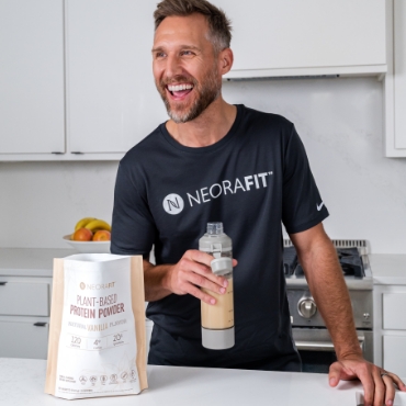 A man wearing a black NeoraFit shirt next to a kitchen counter holding up a glass of water with a bag of Neora's Plant-Based Protein Powder in front of him.